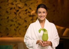 Charming middle aged European woman in white bathrobe, holding a glass with freshly squeezed vitamin juice in her hands, smiles toothy smile looking at camera while enjoying relaxation at wellness spa photo