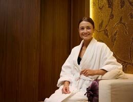 Portrait of a relaxed serene middle aged Caucasian pretty woman in white bathrobe sitting on a couch, smiles toothy smile looking at camera while resting in a luxurious health spa lounge area photo