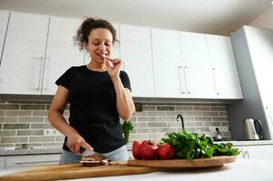 African American woman standing behind a kitchen table,and tasting raw mushroom while chopping ingredients for pizza photo