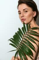 Attractive elegant natural beauty woman in beige underwear posing with exotic palm leaf, standing three quarters against white background with copy space photo