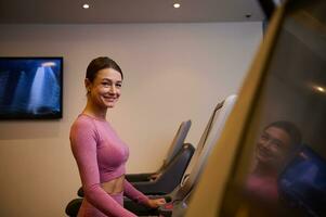 lado ver de un medio Envejecido y alegre mujer en ropa de deporte corriendo en un rueda de andar a gimnasio y sonrisas mirando a cámara. cardio ejercicio. foto