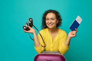 Attractive curly dark-haired beautiful woman smiles toothy smile looking at camera, holding binoculars and passport with boarding pass and ticket, posing with suitcase on blue background photo