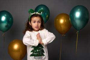 hermosa pequeño niña en Navidad suéter y duende aro poses con manos palma a palmera, mira a cámara, aislado terminado gris antecedentes con dorado metálico verde aire globos con Copiar espacio para anuncio foto