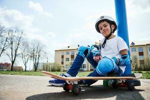 Bottom view of smiling boy in protective gear of skateboarder sitting on skateboard and looking at camera photo