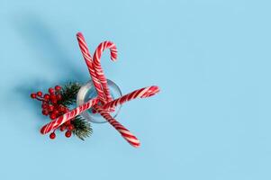 Flat lay of beautiful minimalistic simple composition with Christmas lollipops in transparent glass and snowy branch of pine with red berries, holly, on blue background with copy space for ad photo