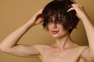 Close-up of a beautiful young Caucasian woman with wet hair, massaging her head looking at camera, isolated over beige background with copy ad space. Hair care and beauty treatment concept photo