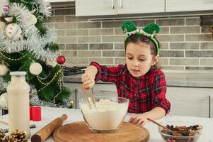 Charming girl with elf hoop on her head thoroughly mixes dry ingredients with flour in glass bowl standing on the table next to the Christmas tree. Little girl cooking, preparing dough at home kitchen photo