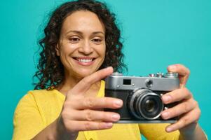 Cheerful African American woman photographer in bright yellow clothes smiling toothy smile while holding an old retro style vintage camera and photographing, taking pictures against blue background photo