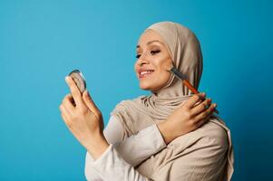 Beautiful Arabic Muslim woman in hijab holds a cosmetic mirror and a makeup brush and applies blush to the cheekbones of her face photo