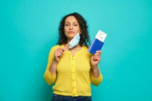 Beautiful mid adult woman traveler in yellow sweater puts her medical mask off and confidently looks at camera, holding a boarding pass, air flight ticket and passport. Copy ad space. Travel concept photo