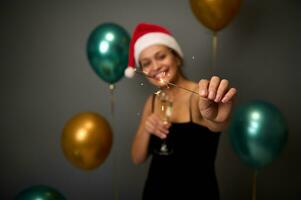 Focus on sparks of sparklers in the hands of a joyful woman having fun at a Christmas or new year party on a gray background with copy space decorated with luxury shiny golden and green balloons photo