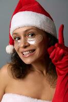Close-up beauty face portrait of attractive young brunette with beautiful smile, wearing Santa hat and smoothing patches under eyes points finger up on copy space on gray background, looking at camera photo