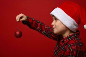 Side close-up portrait of an adorable child boy in a Santa hat holding a red shiny spherical toy of the Christmas tree in front of him, posing on a colored background with copy space for New Year ad photo