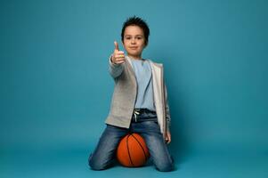 joven baloncesto jugador sentado en un naranja pelota y demostración un pulgar arriba foto