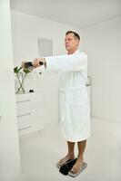 Handsome mature 50 years old man in white terry bathrobe is weighed on a medical smart scale to determine biological age and vitality at a spa wellness center. Health care and medical concept for ad photo