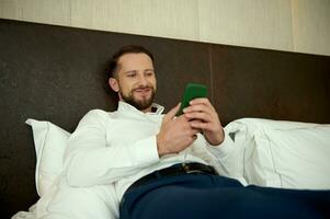 Attractive middle aged European successful businessman, smiles browsing on mobile phone, lying on a bed in the bedchamber of a hotel, relaxing after hard day at work and business meetings photo