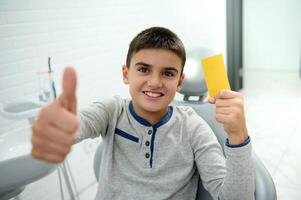 adorable hermoso niño chico sonrisas con hermosa con dientes sonrisa y muestra pulgar arriba y amarillo visitar tarjeta con Copiar anuncio espacio, mirando a cámara mientras sentado en del dentista silla después dental tratamiento foto