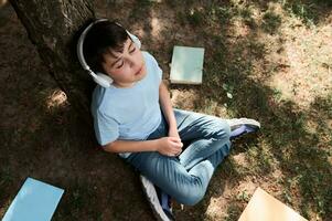 Relaxed teenage boy listens to music on headphones, sitting on the grass near copybooks scattered on the grass in a park photo