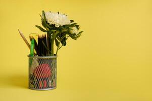 estar para plumas y lapices en el escritorio en un amarillo antecedentes con Copiar espacio. de cerca de un metal Cubeta con papelería. poseedor con de colores colegio suministros y aster flor foto