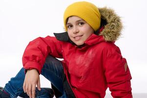 Beautiful child, handsome preadolescent European boy in bright yellow hat and warm red down jacket smiles toothy smile looking at camera sitting on a white background with copy space for advertisement photo