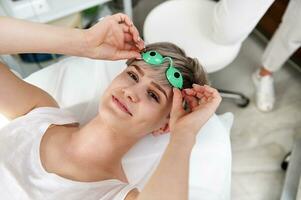 Smiling beautiful client of cosmetology clinic putting off protective UV goggles after laser epilation procedure photo