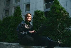 Lifestyle portrait of a young cute woman freelancer, copywriter, motivated start-up sitting on parapet with laptop, working remotely and smiling cute to camera. Connection and online business concept photo