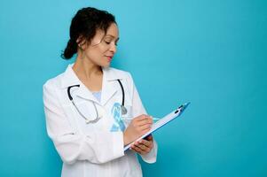 Middle aged mixed race woman, female doctor wearing a blue satin ribbon, symbolic bow color raising awareness in diabetes day , makingtes on a clipboard, isolated over blue background with copy space photo