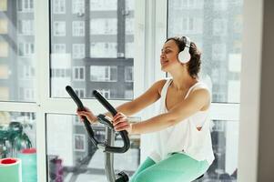 Young attractive woman listen to the music and working out on spin bike against large windows at home photo