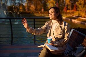 Mixed race pretty woman in beige outfit sitting on a park bench with cup of coffee and hand book, waving hello while looking away on background of beautiful lake with reflected yellow autumn leaves photo