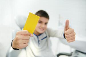 Focus on blank yellow visit card with copy space for insert ads and text in the hand of blurred handsome boy smiling toothy smile and showing thumb up looking at camera sitting in dentist chair photo