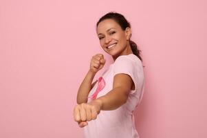 bonito mujer en rosado camiseta y cáncer conciencia cinta soportes en luchando postura a marca el lucha en contra cáncer, en honor de octubre Primero , sonrisas mirando a cámara, de colores fondo, Copiar espacio foto