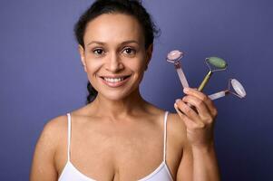 Beauty portrait of an African American pretty woman with vitiligo skin problems, in white underwear smiles with a beautiful toothy smile holding colorful facial roller massagers near her face. photo