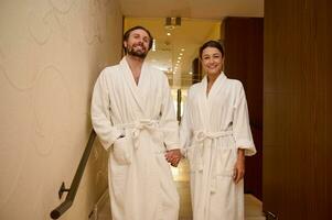 Adorable newlywed couple in love, dressed in white bathrobes, enjoying relaxation together in luxury spa during their honeymoon, holding hands as they walk down the stairs of the wellness spa complex photo