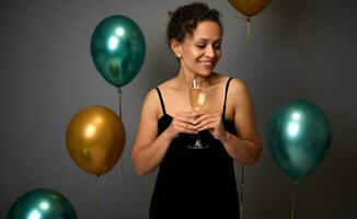 Charming middle aged mixed race woman in evening black velvet dress posing with a glass of champagne, isolated over gray background and beautiful gold and green metallic balloons. Copy space for ad photo