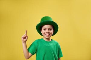 Funny little boy in Irish leprechaun hat pointing his index finger up. Saint Patrick Day on yellow background with copy space photo