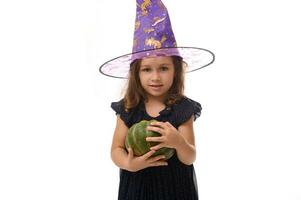 Waist-length shot of little caucasian girl dressed in stylish carnival attire and wizard hat, witch child posing with pumpkin against a white background with copy space. Halloween Witch concept photo