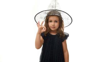Portrait of pretty little witch girl wearing a wizard hat and dressed in stylish carnival dress, gesturing, showing OK sign with fingers. Halloween concept on white background with copy space photo