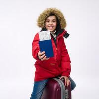 Cheerful preteen boy in bright red jacket with hood sitting on his luggage, smiles toothy smile looking at camera and showing a passport with boarding pass. Winter holidays, travel concept, copy space photo