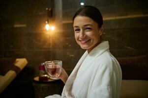 Attractive elegant European young woman in white bathrobe, holding a glass cup of hot healthy herbal drink, looking at camera, sitting on chaise lounge by jacuzzi, enjoying spa procedures in spa salon photo