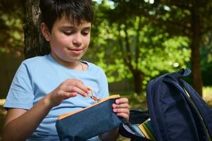 Caucasian teenage boy, smart primary school student opening his pencil case while doing homework in the park after class photo