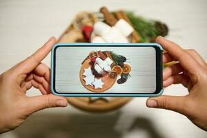 Phone in real time. Close-up. Hands holding phone and taking pictures of gingerbreads in the shape of stars laid out on wooden board and on baking paper next to dried orange slices, pine cones, twigs photo