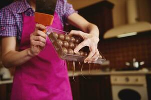 Closeup of chocolatier making shells for chocolate pralines and removing excess chocolate from molds. World Chocolate day concept photo