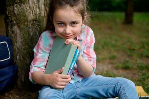 de cerca pequeño chica, inteligente primero grado alumno, primario colegio alumno con libros de texto, lindo sonrisas mirando a cámara foto