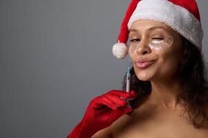 Sexy brunette in Santa hat, wears under eyes patches, attractively looks at camera, holds syringe with beauty injection against gray background. Copy space for Christmas ad. Anti-aging cosmetology photo