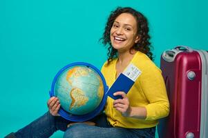 Charming woman traveler in yellow sweater sits next to luggage, holds globe, boarding pass, ticket and passport, smiles toothy smile looking at camera. Tourism concept on blue background, copy space photo
