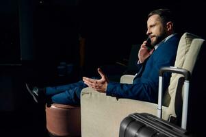 Handsome businessman, business investor sitting relaxed on an armchair with outstretched crossed legs on an ottoman at hotel lobby making a phone call, waiting for someone, during a business trip photo