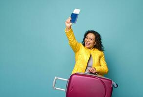 Happy African American woman in yellow clothes with suitcase, passport and ticket with boarding pass, smiled cheerfully posing against blue background. Holidays, travel, vacation and tourism concept photo