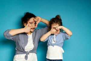 Woman and her baby girl, alike dressed, looking at the camera through their fingers, framing their faces. Blue background with copy space photo