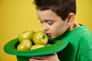 pequeño linda chico disfruta el olor de verde manzanas en un verde irlandesa duende gorra. cara retrato en amarillo antecedentes. Santo patrick's día foto