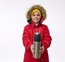 Handsome cheerful school aged boy in bright warm clothes smiles toothy smile looking at camera holding a vacuum flask in front of him, isolated on white background with copy space. Winter trip concept photo